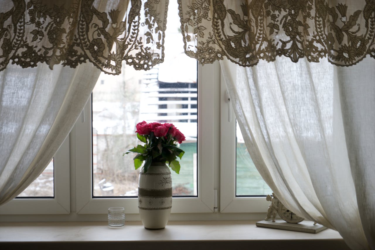 Soft light filters through lace curtains framing a vase of pink roses on a windowsill.